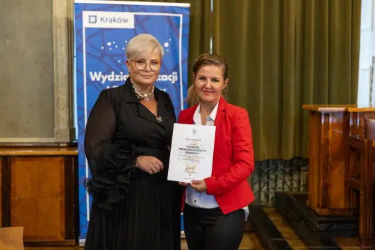 Dwie kobiety trzymają dyplom w sali z zielonymi zasłonami.