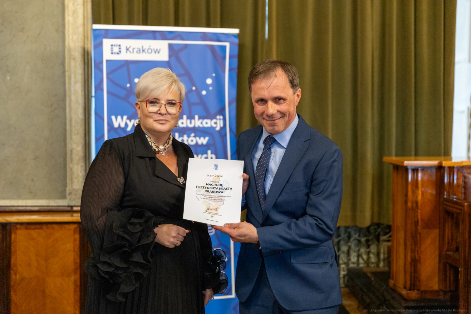 Wręczenie nagrody przez mężczyznę kobiecie, oboje uśmiechnięci, w tle baner z logo Krakowa.  Autor: Bogusław Świerzowski