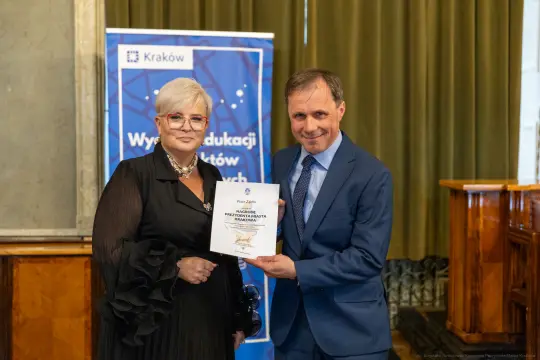 Wręczenie nagrody przez mężczyznę kobiecie, oboje uśmiechnięci, w tle baner z logo Krakowa.