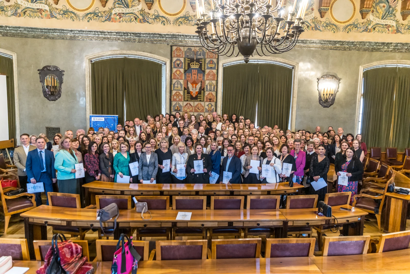 Grupa ludzi w sali, trzymających dyplomy, w tle dekoracyjne ściany i żyrandol.  Autor: Bogusław Świerzowski