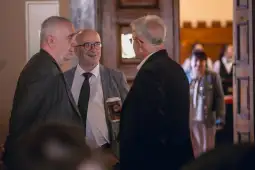 Trzech mężczyzn rozmawia w eleganckim wnętrzu, jeden trzyma kubek z napojem.
