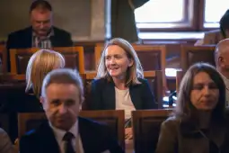Kobieta w jasnej bluzce siedzi w sali obrad, uśmiechając się do innych uczestników.