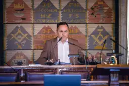 Mężczyzna w garniturze przemawia przy stole w sali obrad, w tle kolorowy gobelin.