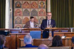 Mężczyzna przemawia z mównicy w sali obrad, w tle inna osoba przy stole.