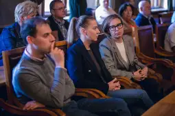 Grupa ludzi siedząca w sali, skupiona na wydarzeniu.