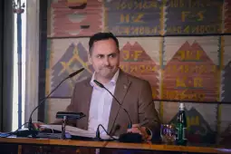 Mężczyzna w garniturze siedzi przy stole, prowadząc dyskusję, z mikrofonem i butelką wody obok.