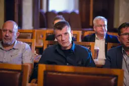 Grupa mężczyzn siedzących w sali, niektórzy z poważnymi wyrazami twarzy.