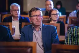 Mężczyzna w okularach siedzący wśród innych uczestników spotkania.