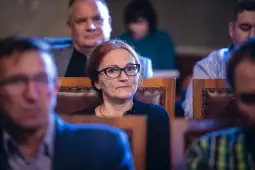 Kobieta w okularach siedząca wśród innych osób w sali.