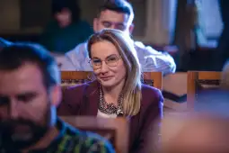 Kobieta w okularach uśmiecha się, siedząc wśród innych uczestników spotkania.