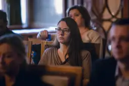 Kobieta w okularach siedząca wśród innych osób, z poważnym wyrazem twarzy.