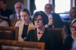 Kobieta w okularach siedząca wśród innych osób w sali.