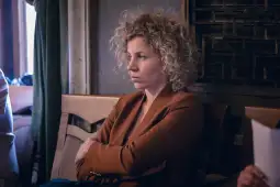 Kobieta z kręconymi włosami w brązowej marynarce, z zamyśloną miną, siedzi na krześle.