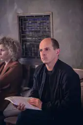 Mężczyzna z kartkami w ręku, siedzący obok kobiety z kręconymi włosami.