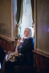 Kobieta z siwymi włosami siedząca w eleganckim wnętrzu, z założonymi rękami.