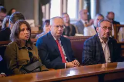 Trzy osoby siedzące w sali, skupione na wydarzeniu, w formalnych ubraniach.
