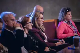 Grupa osób siedzących w sali, skupionych na wystąpieniu.
