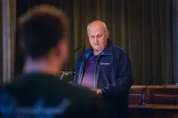 Mężczyzna w niebieskiej kurtce przemawia do publiczności.