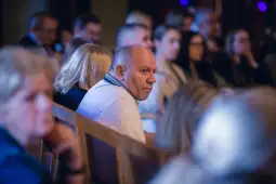 Mężczyzna w białej koszuli siedzi wśród publiczności, skupiony na wydarzeniu.