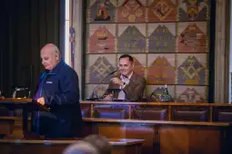 Dwóch mężczyzn w sali obrad, jeden przemawia, drugi słucha i uśmiecha się.