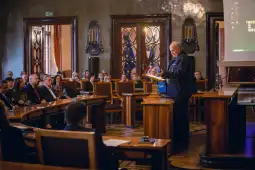 Mężczyzna przemawia do zgromadzonej publiczności w sali konferencyjnej.