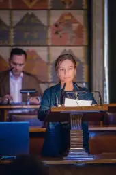 Kobieta przemawia z mównicy, w tle mężczyzna przy biurku.