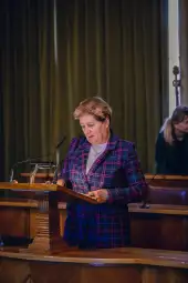 Kobieta w garniturze w kratę przemawia z mównicy w sali obrad.
