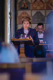 Kobieta w eleganckim stroju przemawia z mównicy, w tle mężczyzna przy stole.