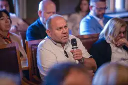 Mężczyzna z mikrofonem zadaje pytanie podczas spotkania, w tle uczestnicy.