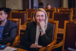 Kobieta w garniturze siedzi w sali obrad, uśmiechając się i myśląc.