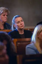 Kobieta w czarnym żakiecie, skupiona, siedzi wśród innych osób w tle.