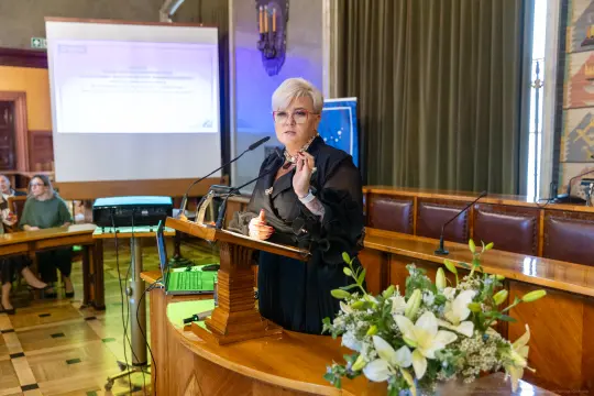 Kobieta przemawia z mównicy podczas konferencji, w tle ekran z prezentacją.