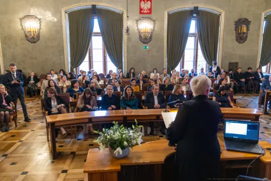 Konferencja z uczestnikami siedzącymi w sali, prelegentka przemawia do zgromadzonych.