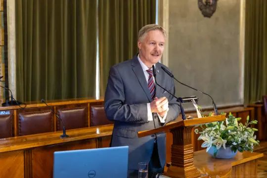 Mężczyzna w garniturze przemawia z mównicy, obok kwiaty i laptop.