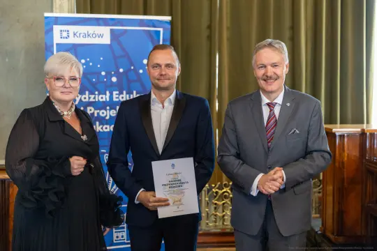 Trzy osoby w formalnych strojach, trzymają dokument, w tle baner z logo Krakowa.