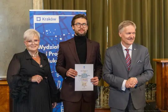 Mężczyzna z dyplomem stoi między dwiema osobami w eleganckich strojach.