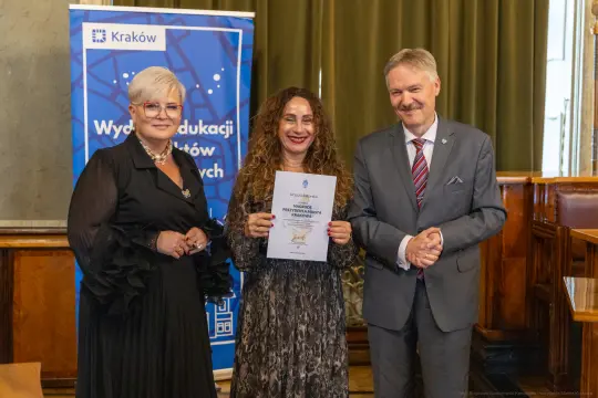 Trzy osoby trzymają dyplom w sali z dekoracjami. W tle logo Krakowa.