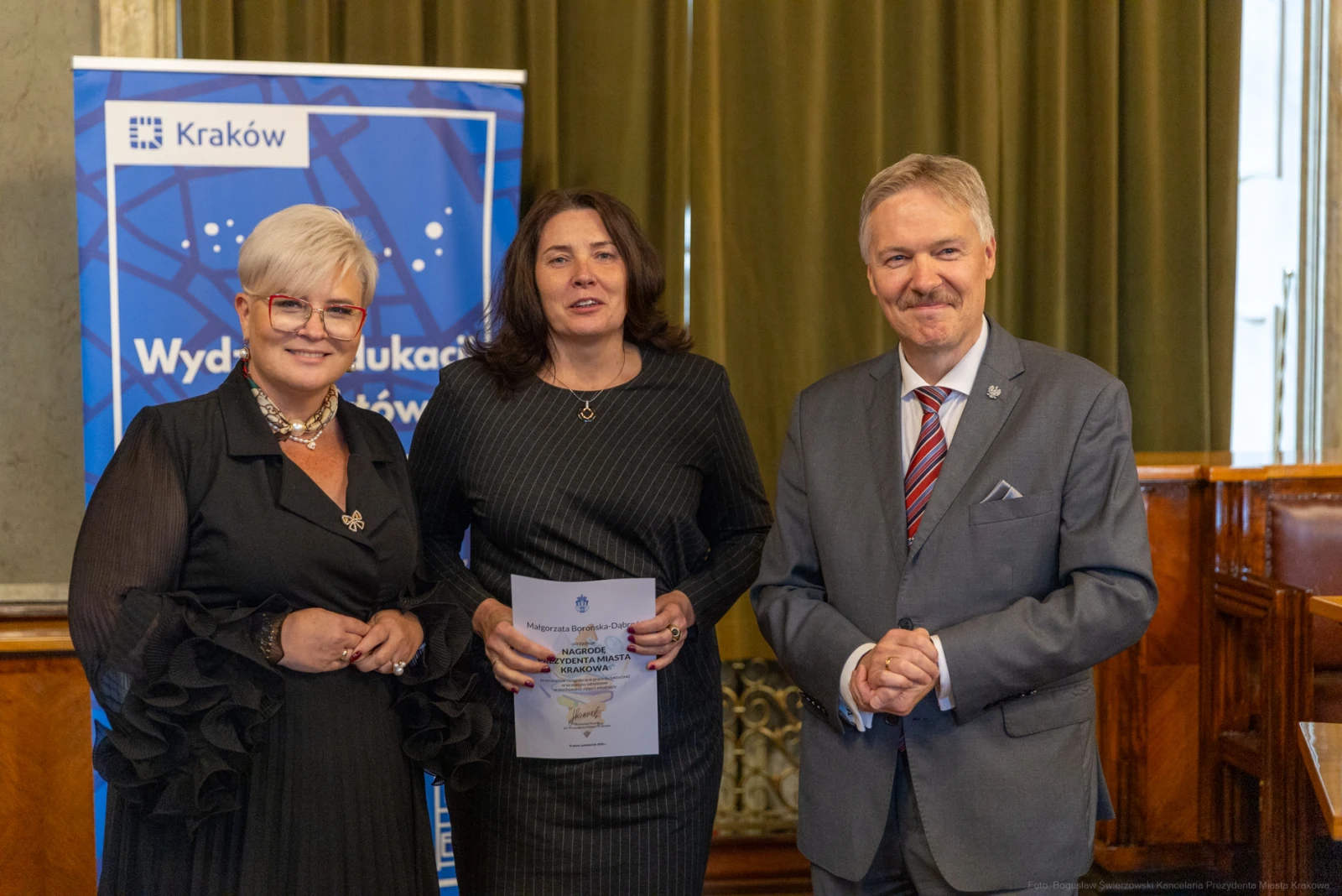 Trzy osoby w formalnych strojach, trzymające dokumenty, w sali z zielonymi zasłonami.  Autor: Bogusław Świerzowski