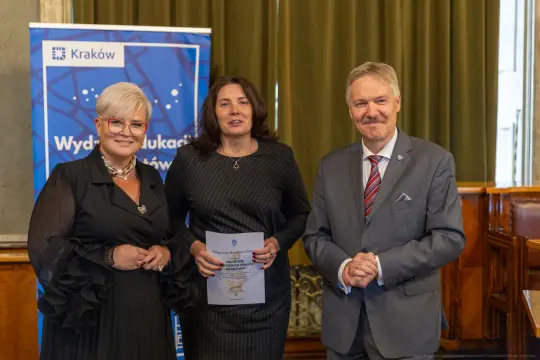 Trzy osoby w formalnych strojach, trzymające dokumenty, w sali z zielonymi zasłonami.