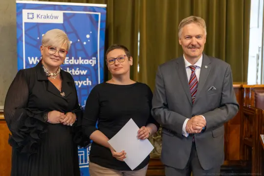 Trzy osoby w formalnych strojach, uśmiechające się, stoją w sali z zielonymi zasłonami.