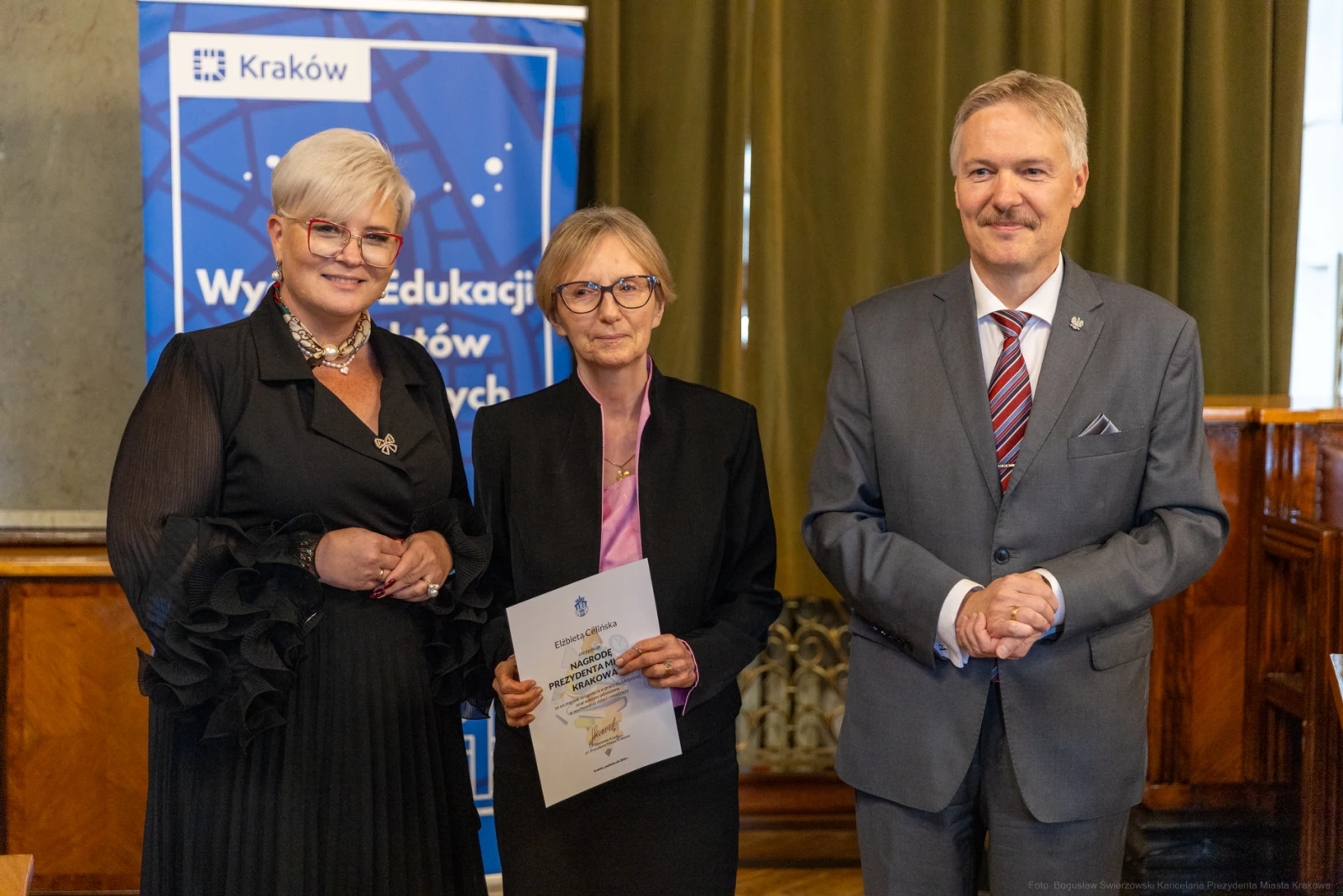 Trzy osoby w formalnych strojach, jedna z dyplomem, w tle baner z logo Krakowa.  Autor: Bogusław Świerzowski