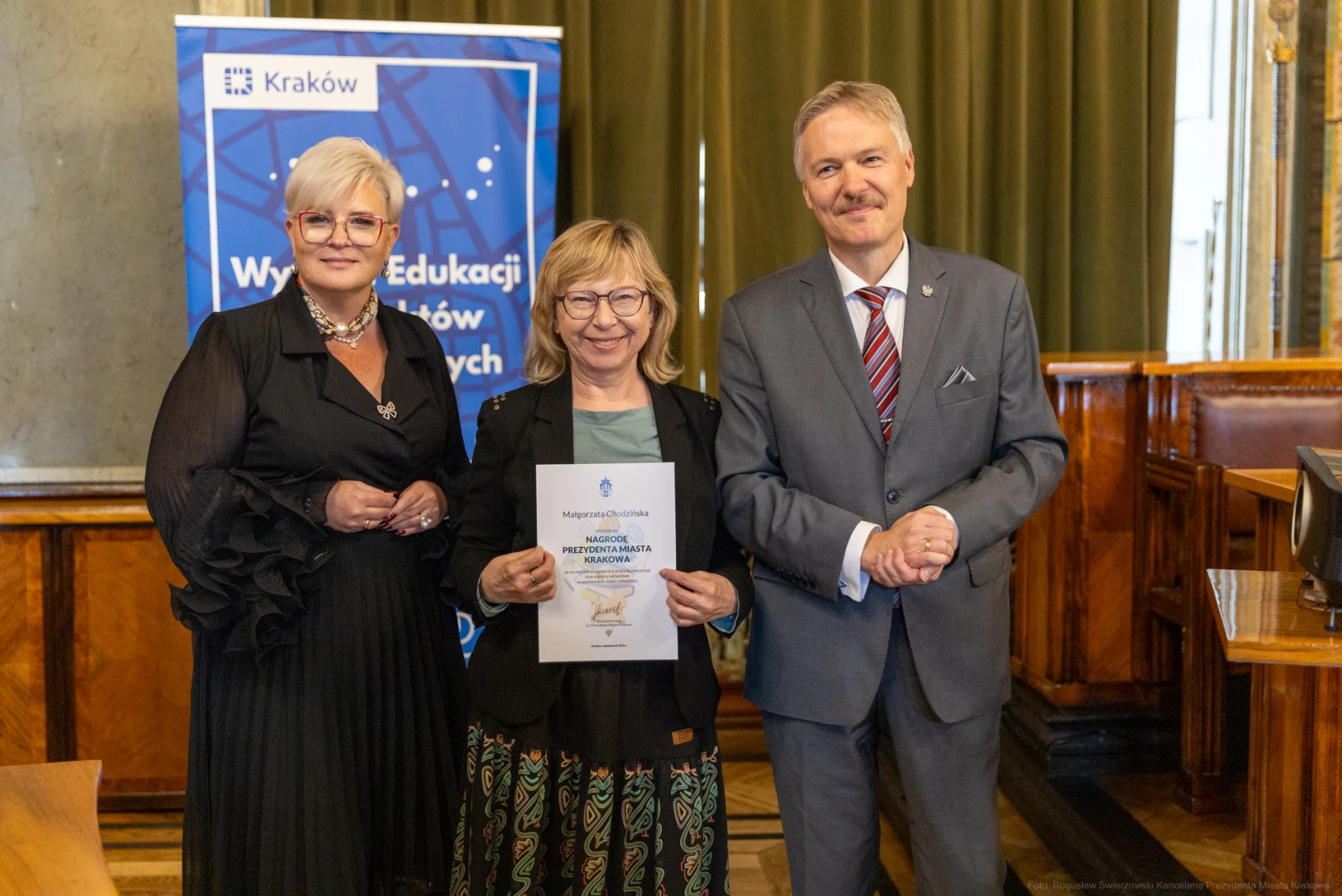 Trzy osoby w formalnych strojach trzymają dyplom w sali z drewnianymi elementami.  Autor: Bogusław Świerzowski