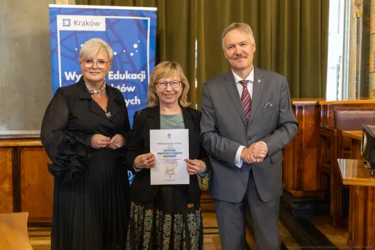 Trzy osoby w formalnych strojach trzymają dyplom w sali z drewnianymi elementami.