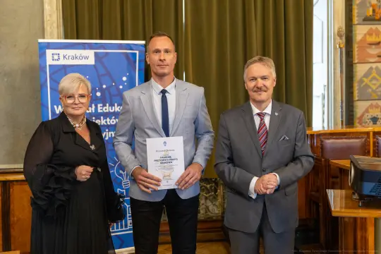 Trzy osoby w formalnych strojach trzymają dyplom w sali z dekoracjami.