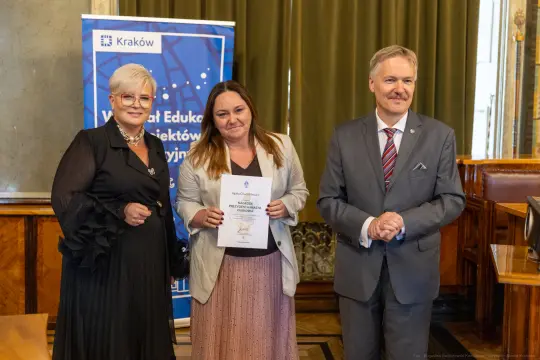 Uroczystość wręczenia nagrody w Krakowie, trzy osoby z dyplomem i banerem w tle.
