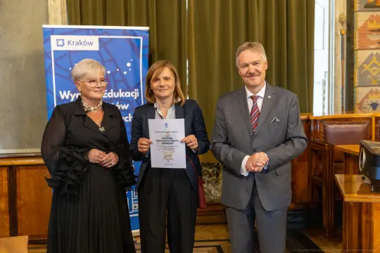 Trzy osoby w formalnych strojach trzymają dyplom w sali z dekoracjami.