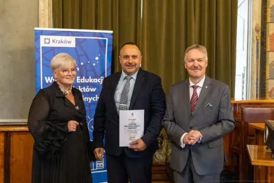 Trzy osoby w formalnych strojach, trzymające dokumenty, w tle baner promujący edukację w Krakowie.