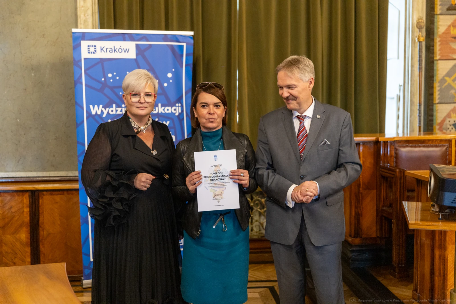 Wręczenie dyplomu w Wydziale Edukacji w Krakowie. Trzy osoby w formalnych strojach.  Autor: Bogusław Świerzowski