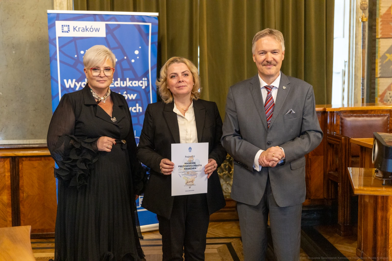 Trzy osoby w formalnych strojach trzymają dokument w sali z zielonymi zasłonami.  Autor: Bogusław Świerzowski