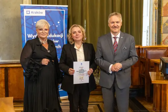 Trzy osoby w formalnych strojach trzymają dokument w sali z zielonymi zasłonami.